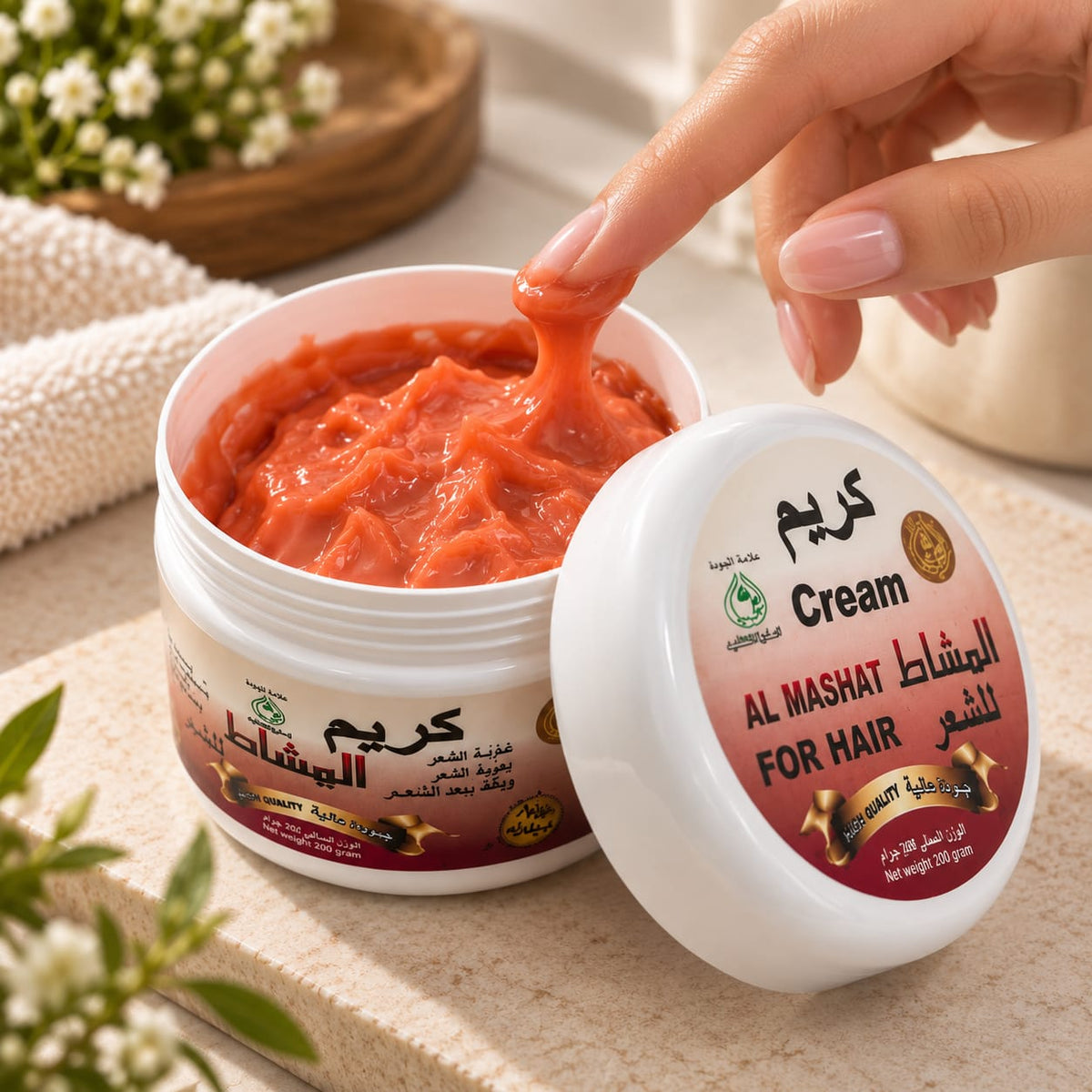 An open white jar of hair cream showing a thick orange-red paste, with a hand lifting some of the cream. The lid sits beside the jar on a beige surface, with Arabic and English text on the label reading 'Cream' and 'AL MASHAT FOR HAIR'.