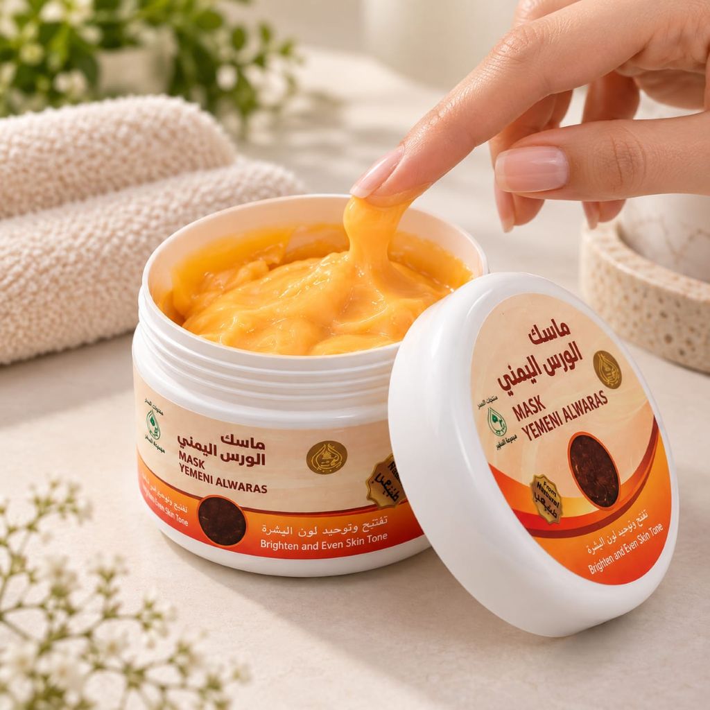 A white plastic jar of Yemeni Al waras Mask Brighten and Even Skin Taze mask, shown open with a hand lifting a stretchy orange mask. The lid is placed beside the jar, showing the product label.