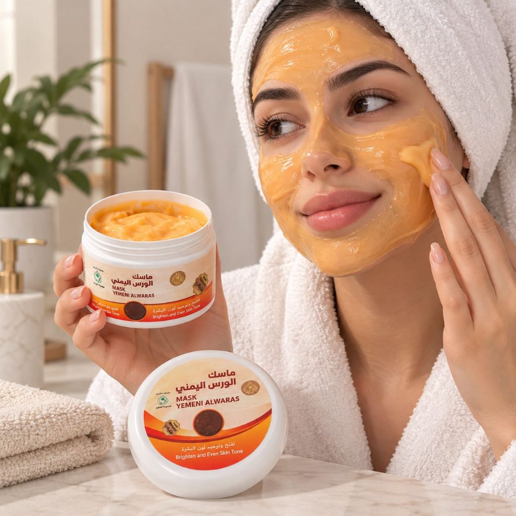 A woman in a white robe applies a Yemeni Al Waras Mask while holding an open jar and a closed container of Yemeni Al Waras Mask Cream. The product packaging features white, orange, and brown colors with Arabic text.