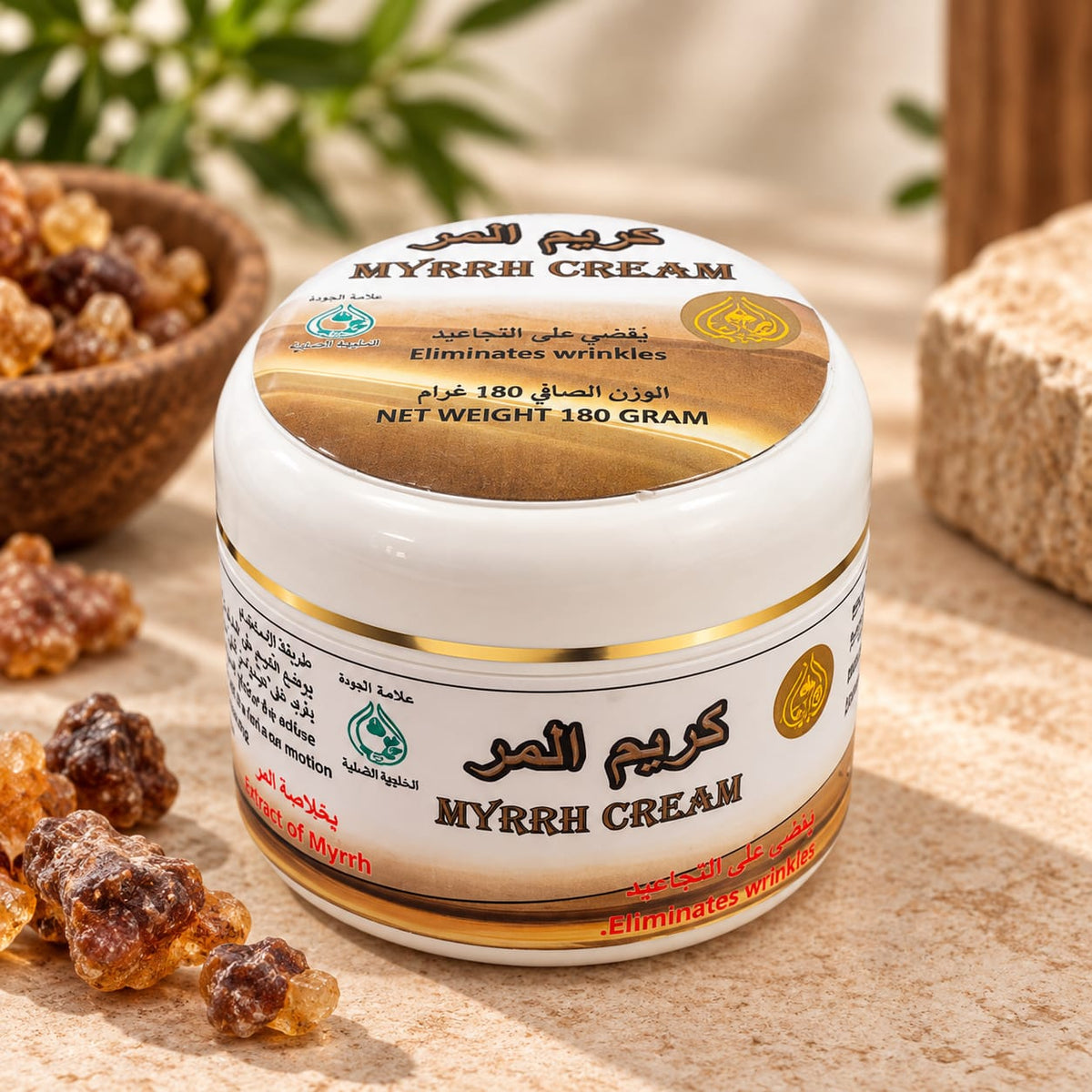 A jar of MYRRH CREAM with Arabic and English text, featuring a white and gold label. The jar is styled on a beige stone surface with raw myrrh resin pieces and a wooden bowl in the background.