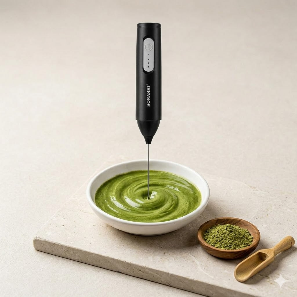 Black handheld milk frother / Powder Mixerin a bowl of green sidr smoothie on a light surface with a bowl of matcha and wooden spoon.