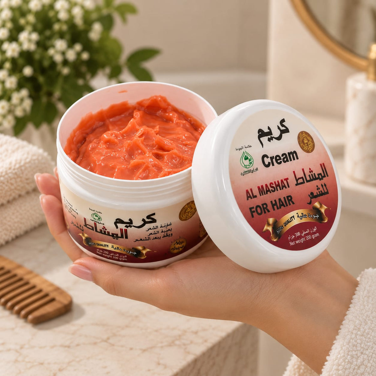 A hand holds an open white jar of Al Mashat Cream for hair, showing the bright orange-red cream inside, with the matching lid placed beside it on a marble surface.