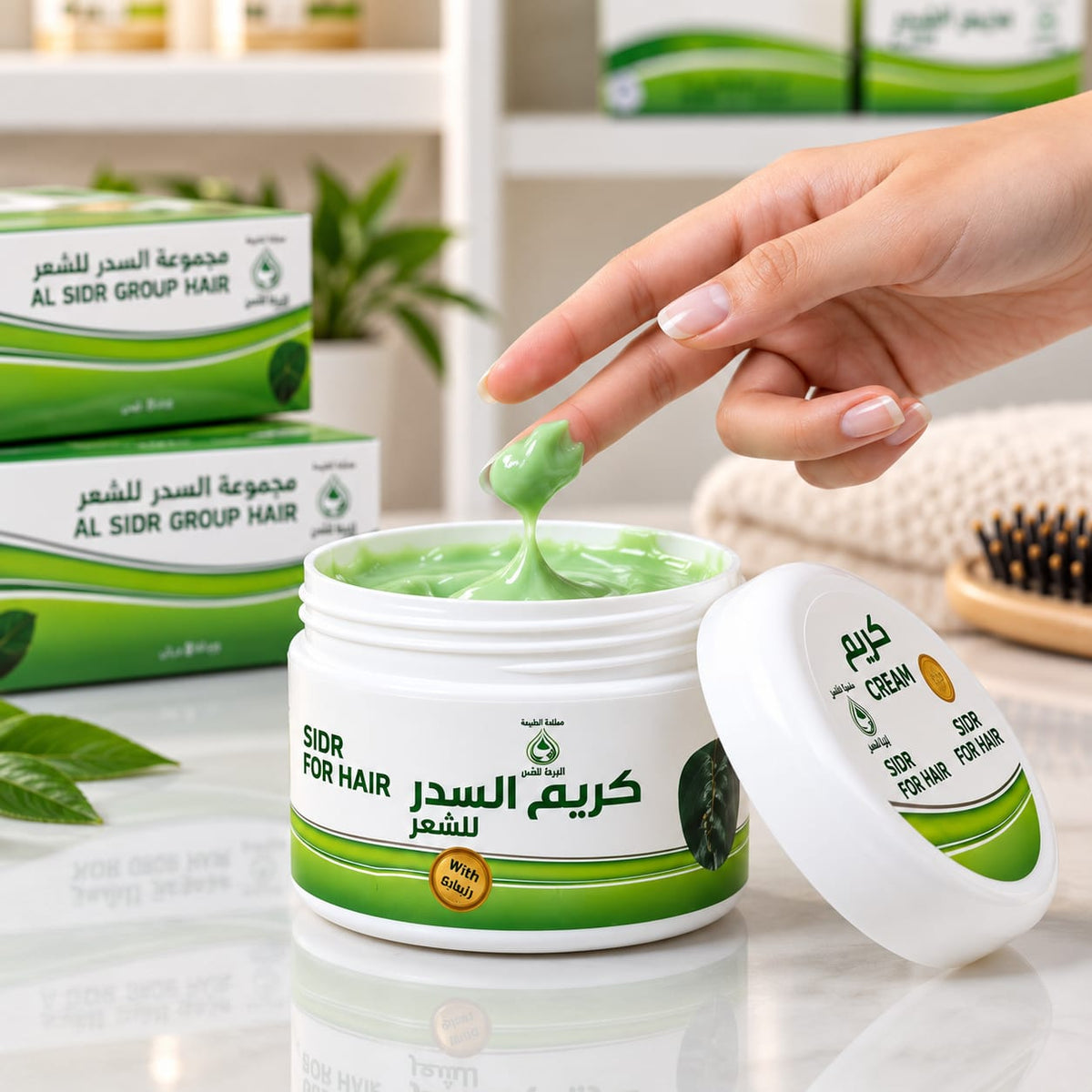 A hand with manicured nails scoops light Al Sidr Cream for hair from an open white jar, with the lid resting beside it. Two branded product boxes are stacked in the background on a white shelf, along with a towel and a hairbrush in a bright indoor setting.