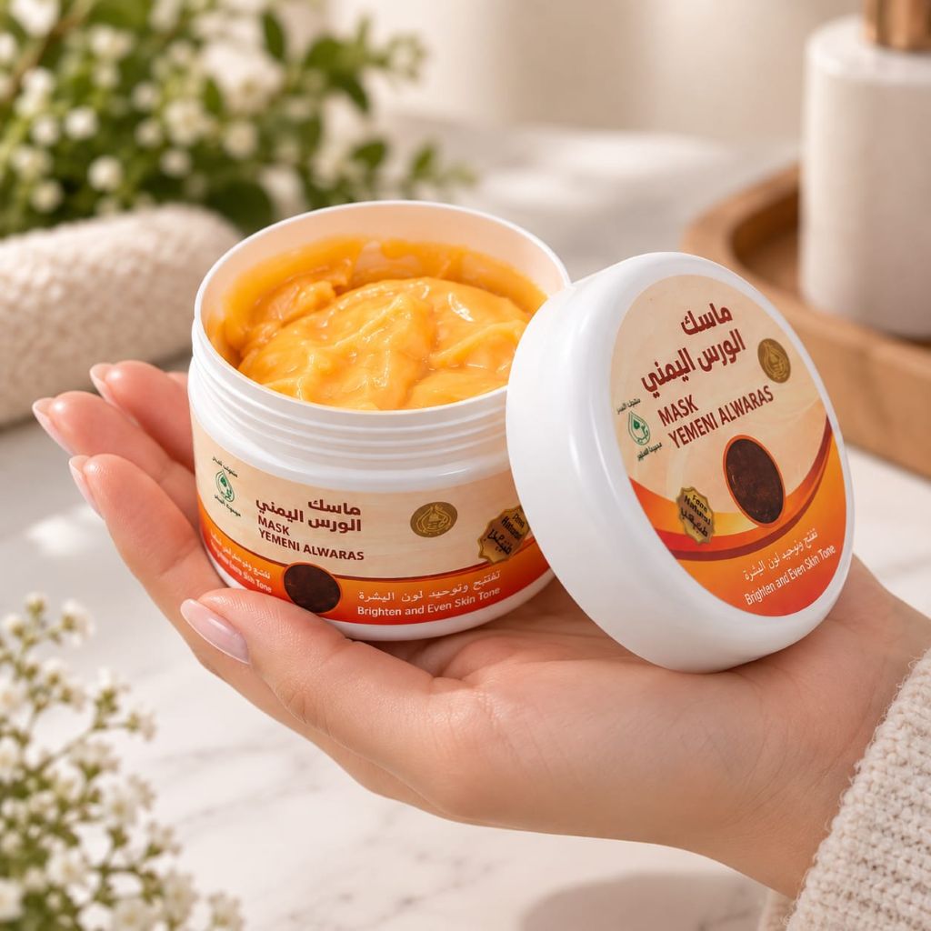 An open white jar of Yemeni Al Waras Mask with orange cream visible inside, held in a person's hand. The matching lid sits beside it on a soft, blurred bathroom-like surface with flowers and a towel in the background.