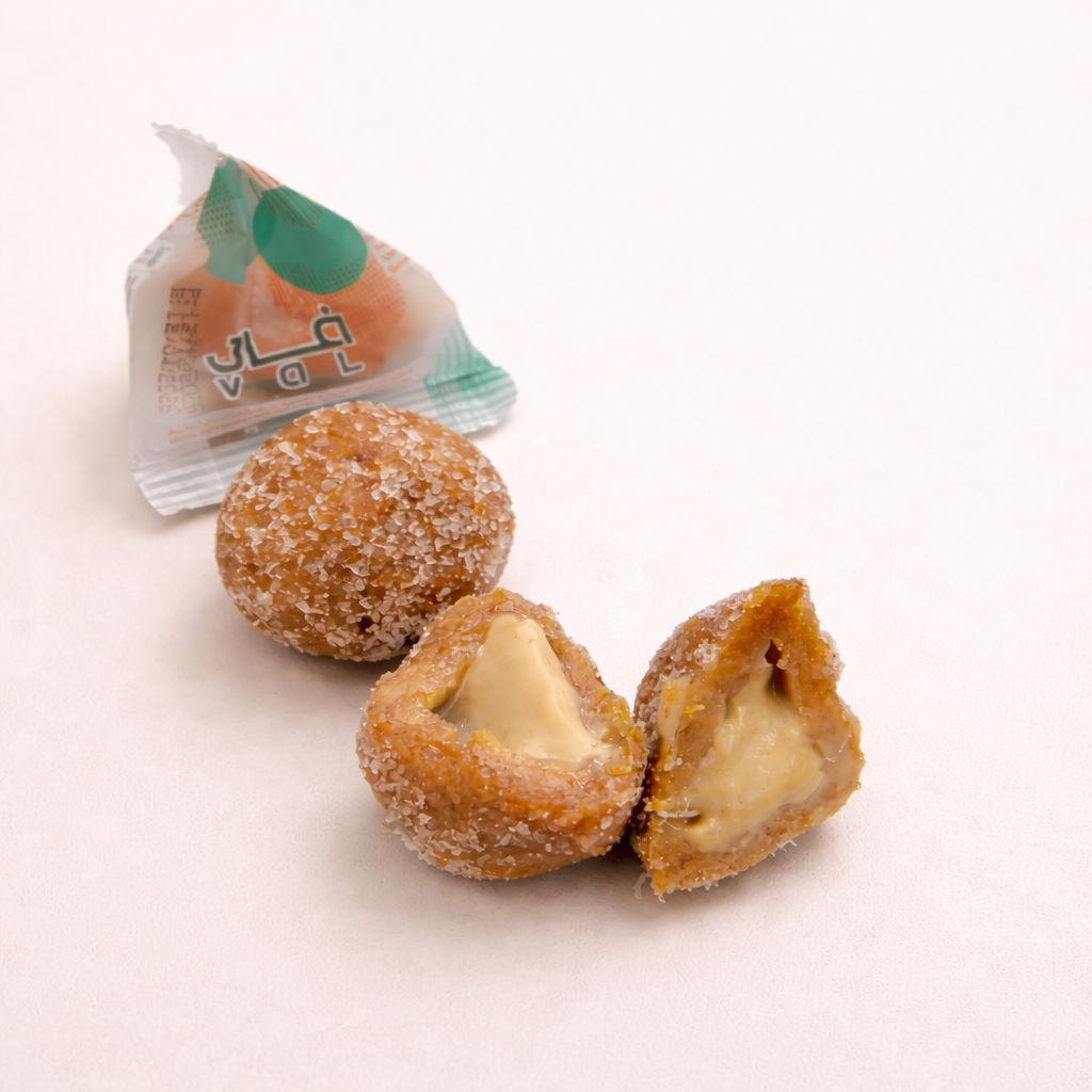 Two half rounded dates stuffed with tahini and coated with coconut, next to a small green and white package on a light background.